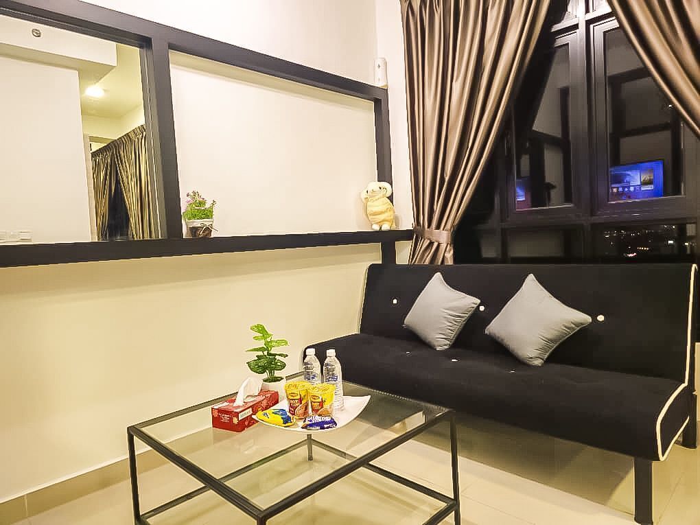 A living room with a black sofa, a glass coffee table with snacks and a plant, a window with curtains, and a TV in the background.