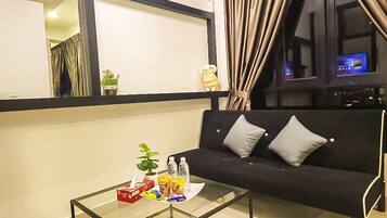 A living room with a black sofa, a glass coffee table with snacks and a plant, a window with curtains, and a TV in the background.