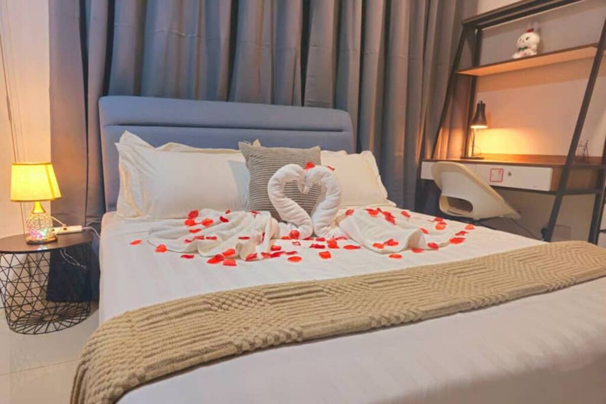 Una cama bien tendida con una toalla dispuesta en forma de corazón, una lámpara de noche y una mesita de noche con lámpara.