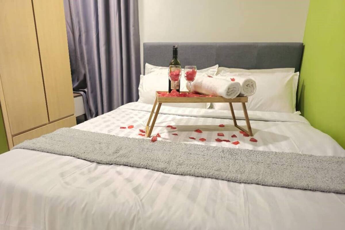 Una cama impecablemente hecha con un edredón gris, una bandeja con una botella y vasos, y pétalos de rosa esparcidos sobre la cama.
