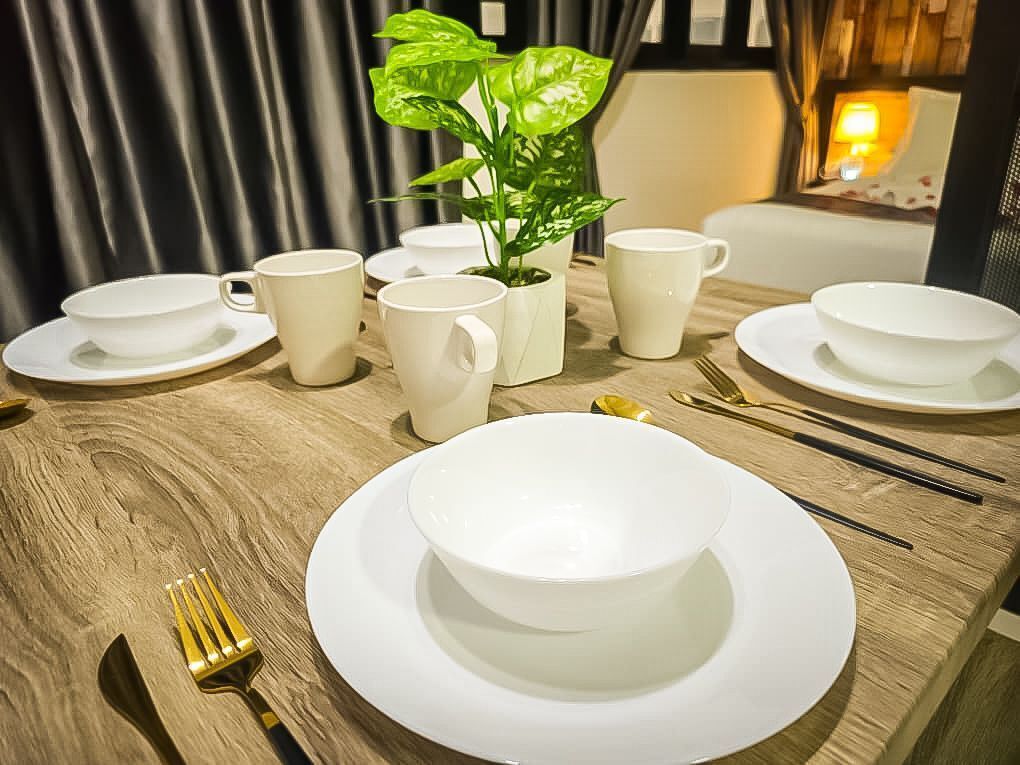A dining table set with white plates, bowls, and cups, a potted plant, and a window with curtains.
