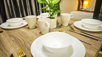 A dining table set with white plates, bowls, and cups, a potted plant, and a window with curtains.