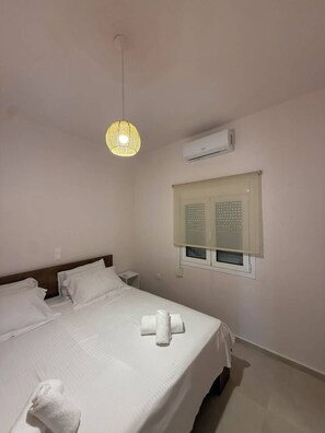 Habitación de hotel con cama, lámpara colgante, equipo de aire acondicionado y ventana con persianas.