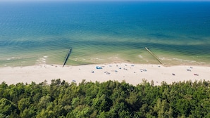 Eine Vogelperspektive zeigt einen Strand mit sandigem Ufer, klarem blauen Wasser und einer bewaldeten Fläche im Vordergrund.