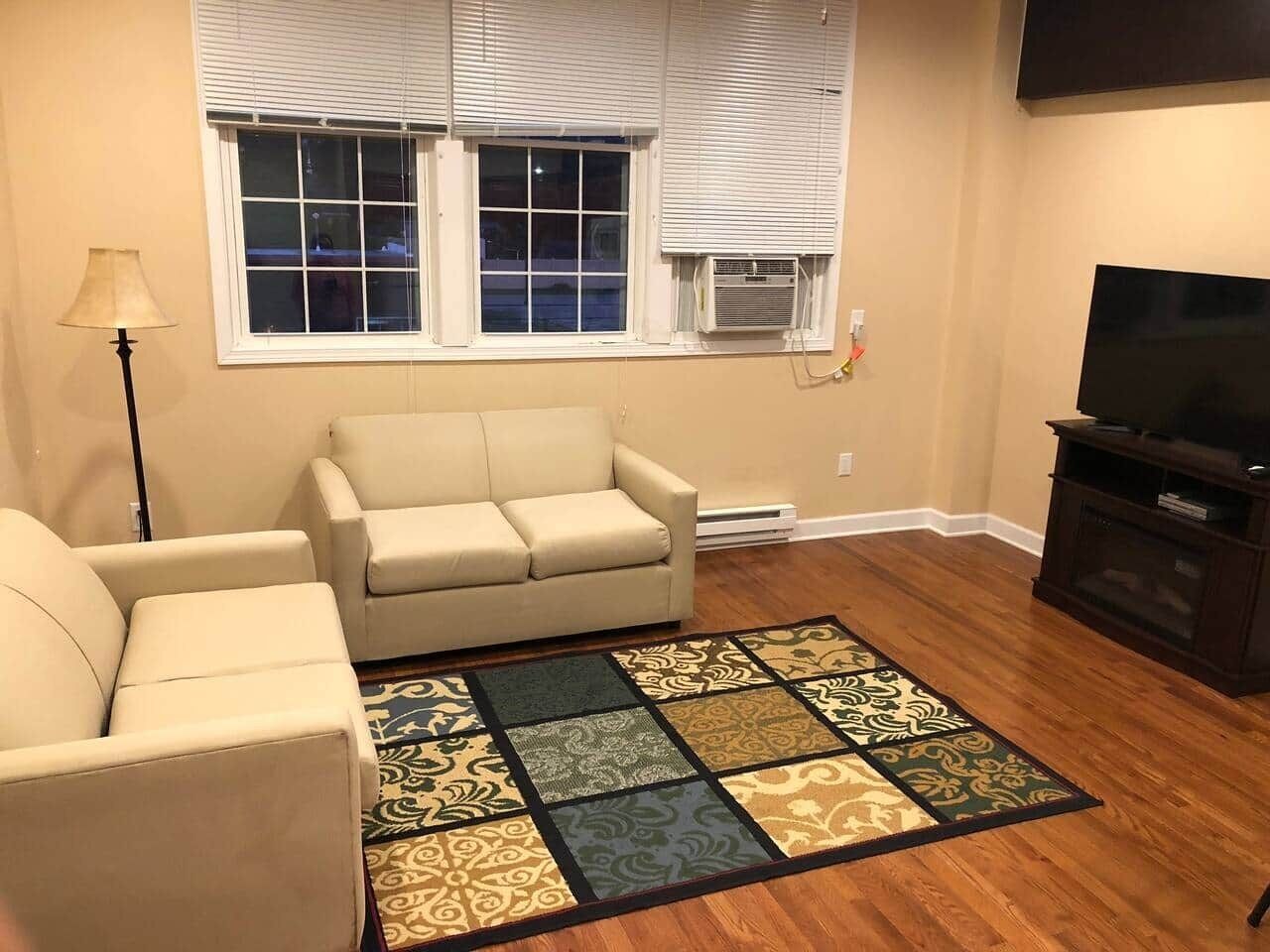 A living room with a beige sofa, a patterned rug, a television, and a window with blinds.