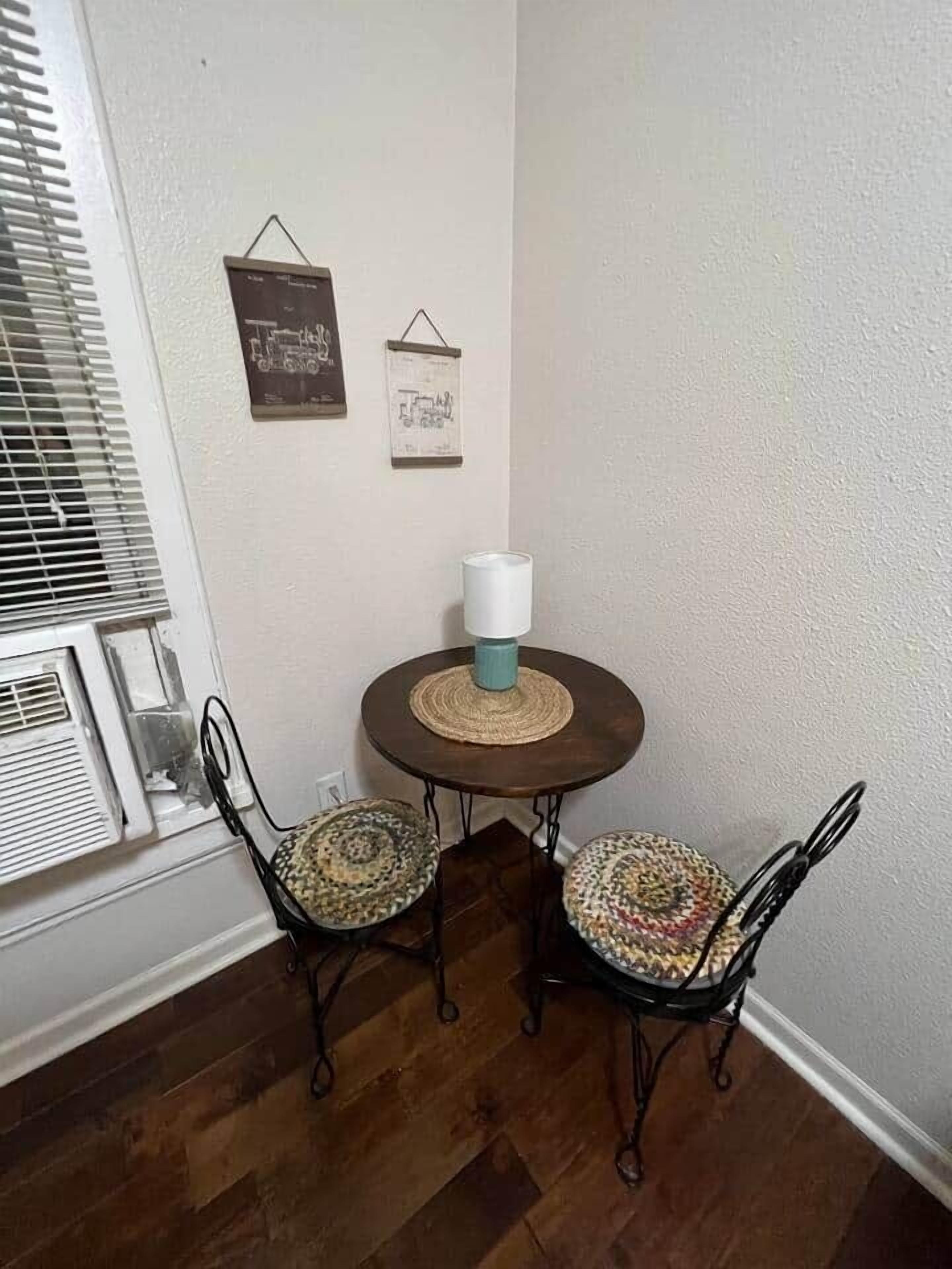 Quarto de canto com uma mesa redonda, duas cadeiras, ar condicionado fixado na parede e dois objetos emoldurados na parede.