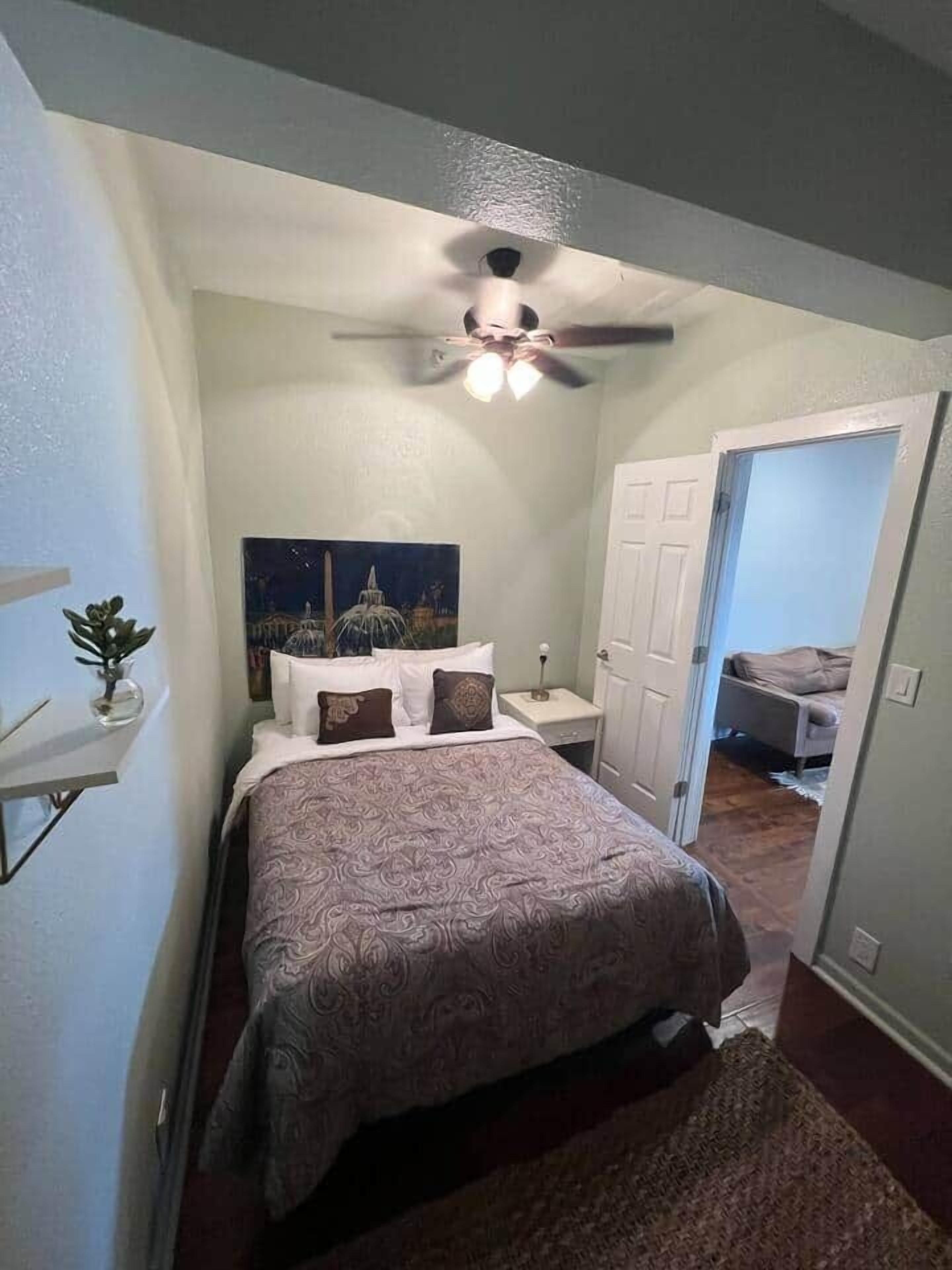 Ein Schlafzimmer mit einem Bett, Deckenventilator, Nachttisch, einem Bild und einer Tür zu einem anderen Raum.