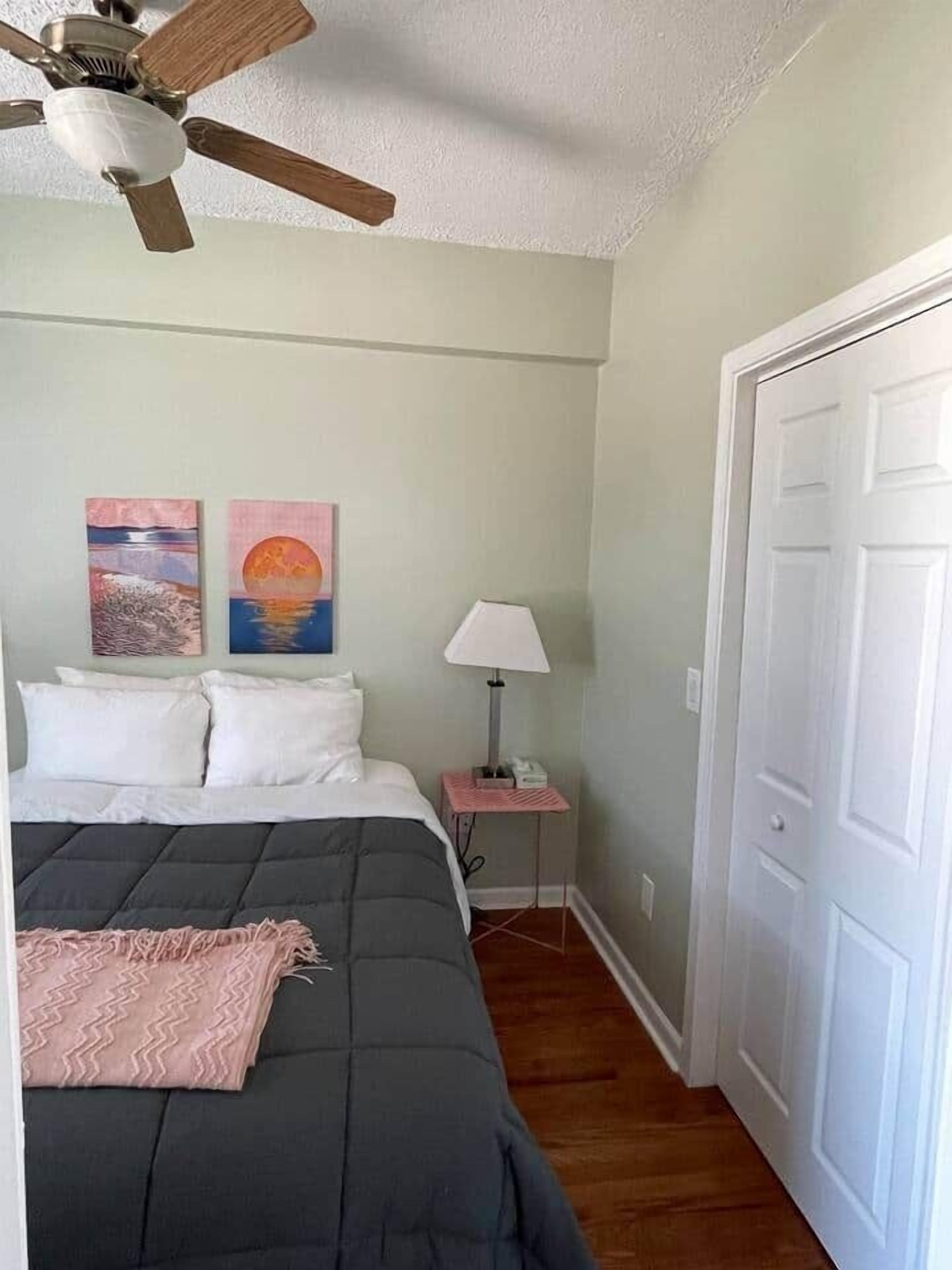 Un dormitorio con cama, ventilador de techo, mesita de noche con lámpara y dos cuadros en la pared.