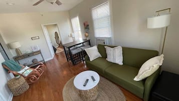 Ein Wohnzimmer mit einem grünen Sofa, einem runden Couchtisch, Korbstühlen, einem Deckenventilator und einem Essbereich mit Tisch und Stühlen.