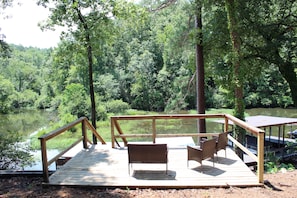 Terraza de madera con dos sillas de mimbre y barandilla, con vista a un lago rodeado de árboles.