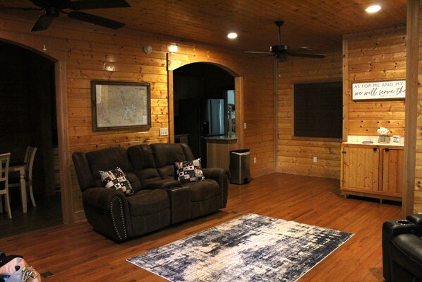 A cozy living room with a dark brown leather sofa, a patterned rug, wooden walls, a ceiling fan, and a kitchen area with a sign on the wall.