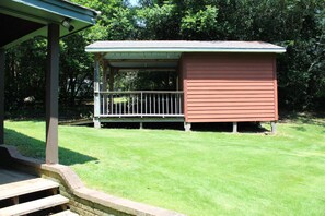 Une petite construction à un seul étage, dotée d’un porche et d’une terrasse délimitée par une clôture, entourée d’une pelouse bien entretenue et d’arbres touffus.