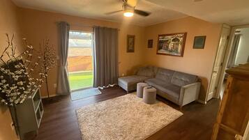 Un salon avec un canapé gris, un petit tapis, un ventilateur de plafond et une vue dégagée sur l’extérieur grâce à une grande fenêtre.