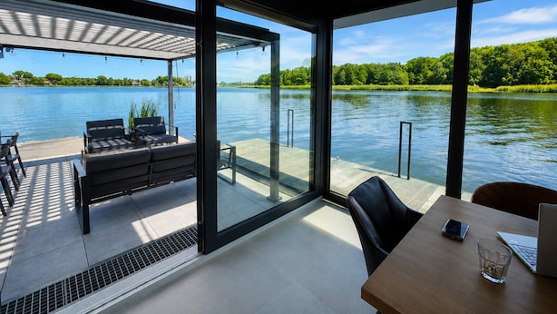 A modern room with a large window overlooking a lake, a wooden table, and a sofa. - Comfortable Dziwnow House for 8 (Dziwnów)