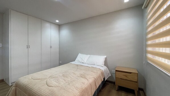 Une chambre à coucher comprenant un lit, une table de chevet en bois, une armoire et une fenêtre avec des stores.