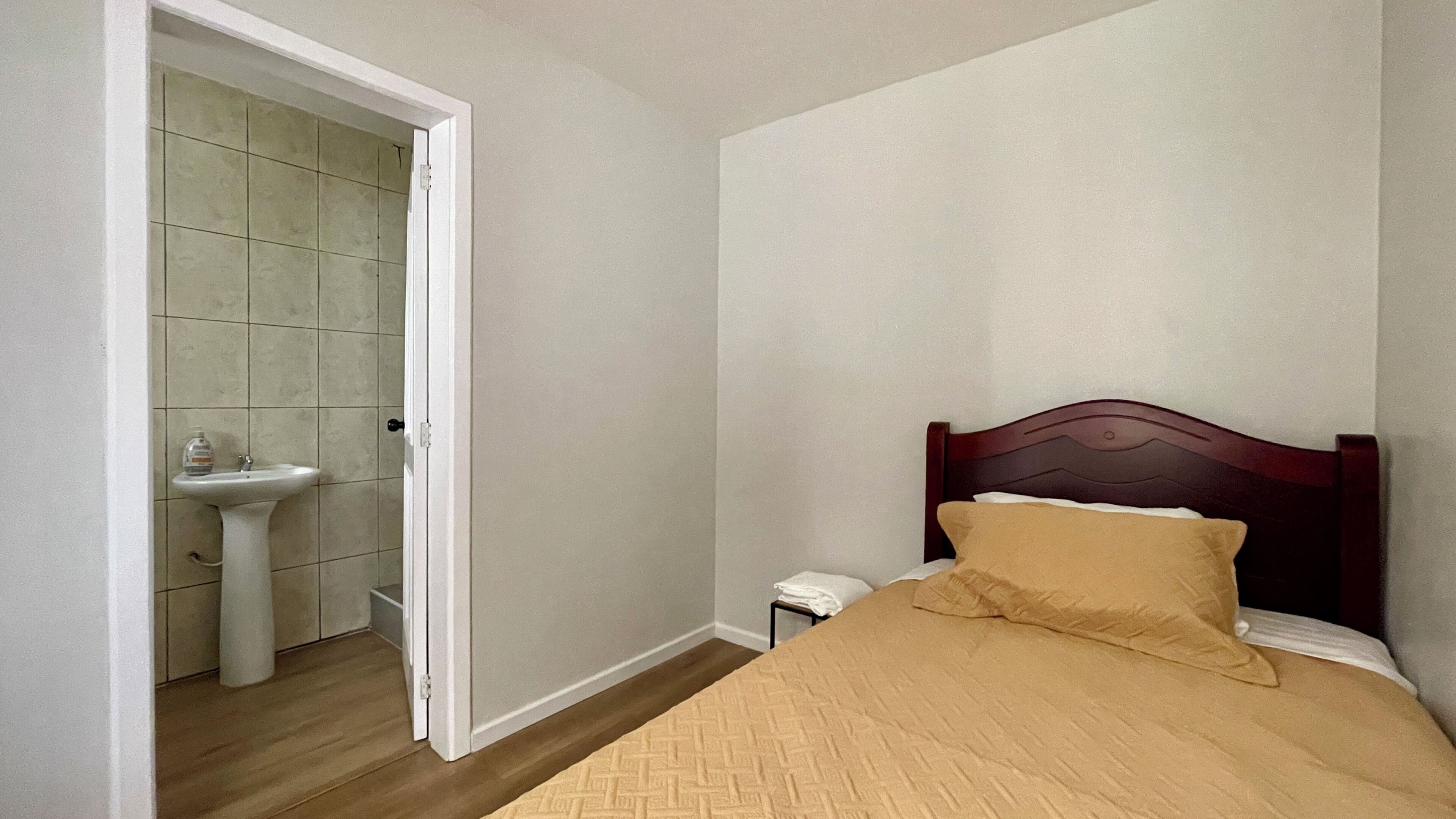 A bedroom with a wooden bed, a bedside table, and a bathroom with a sink and toilet.