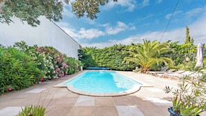 Una piscina rectangular con solárium de baldosas, rodeada de plantas y una pared blanca.