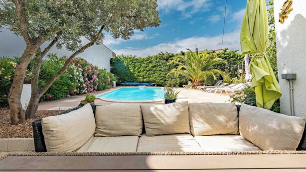 Zona de piscina con un sofá, una mesa techada y una sombrilla verde.
