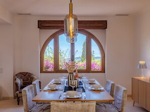 A dining area with a table set for four, a vase with dried flowers, and a view through large arched windows. - Floralies by Interhome (Sainte Maxime)