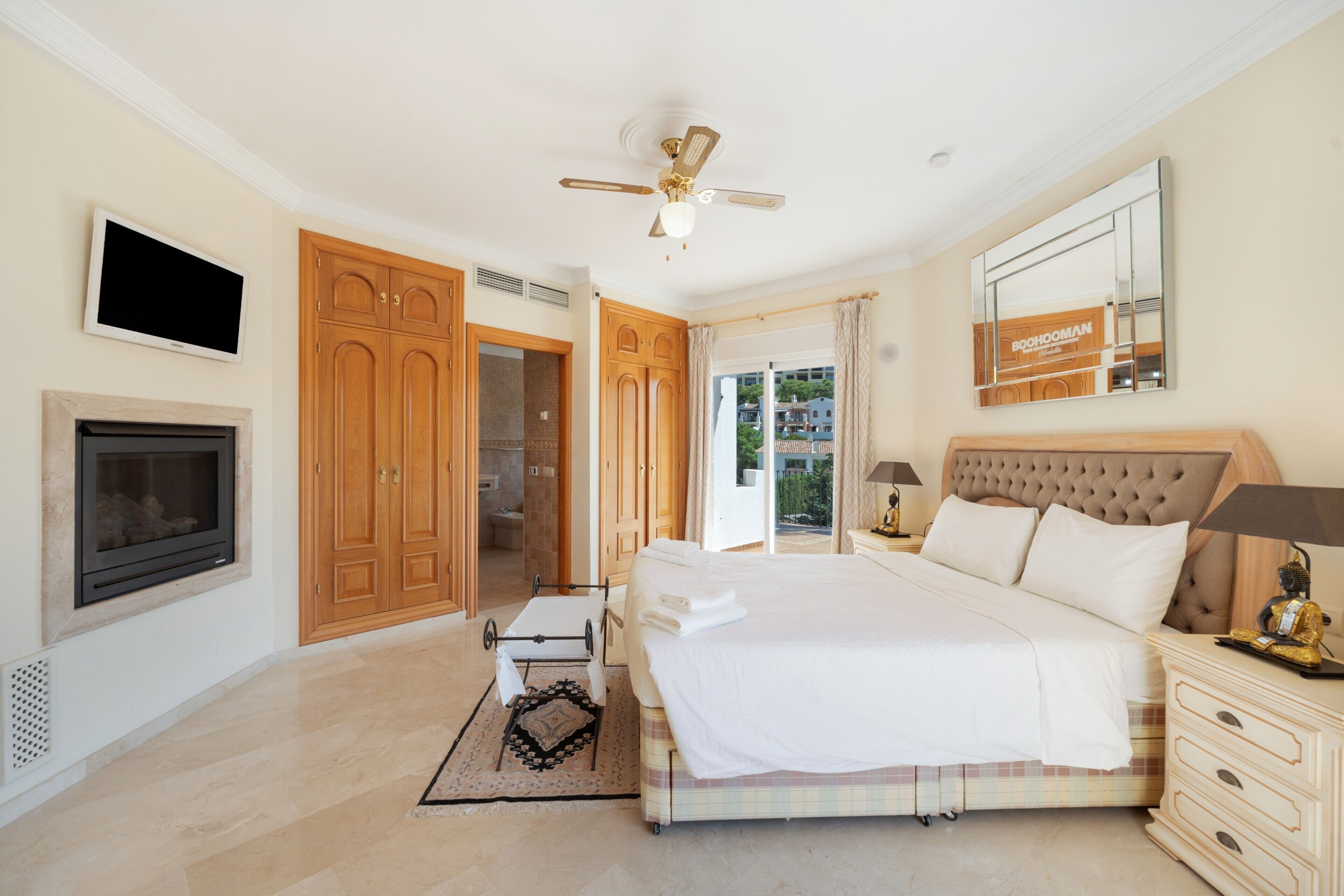 Une chambre à coucher avec un lit, un ventilateur de plafond, une télévision, une cheminée, des portes en bois et une vue par la fenêtre.