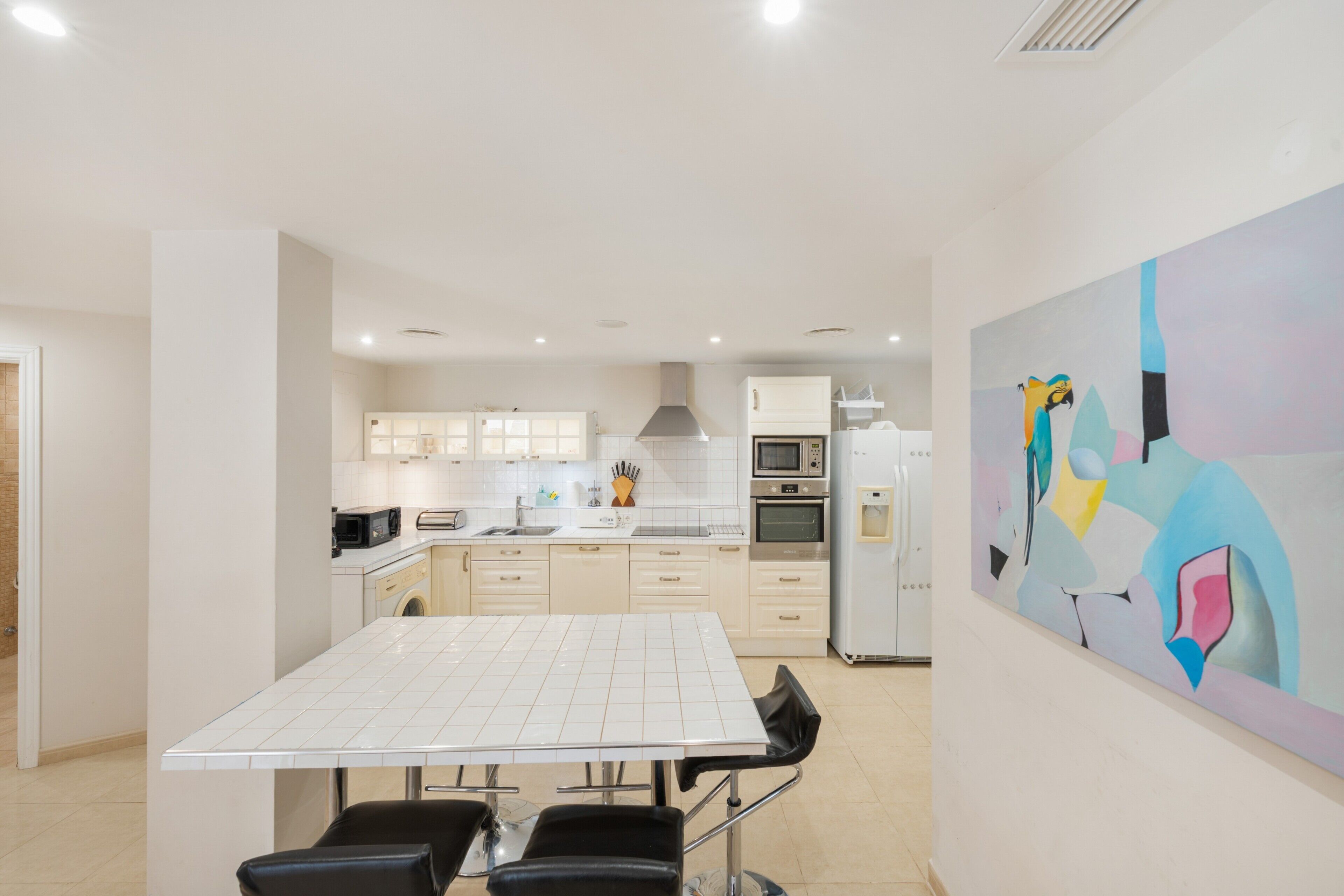 Una cocina moderna con armarios blancos, una mesa de comedor con sillas y un cuadro abstracto de colores en la pared.