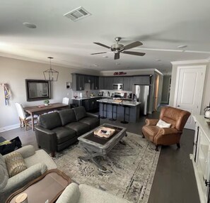 A living room with a sofa, armchair, and coffee table. There is a kitchen area with a bar and a dining table.