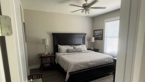 Quarto com cama, mesa de cabeceira, cadeira, ventilador de teto e janela com persianas.