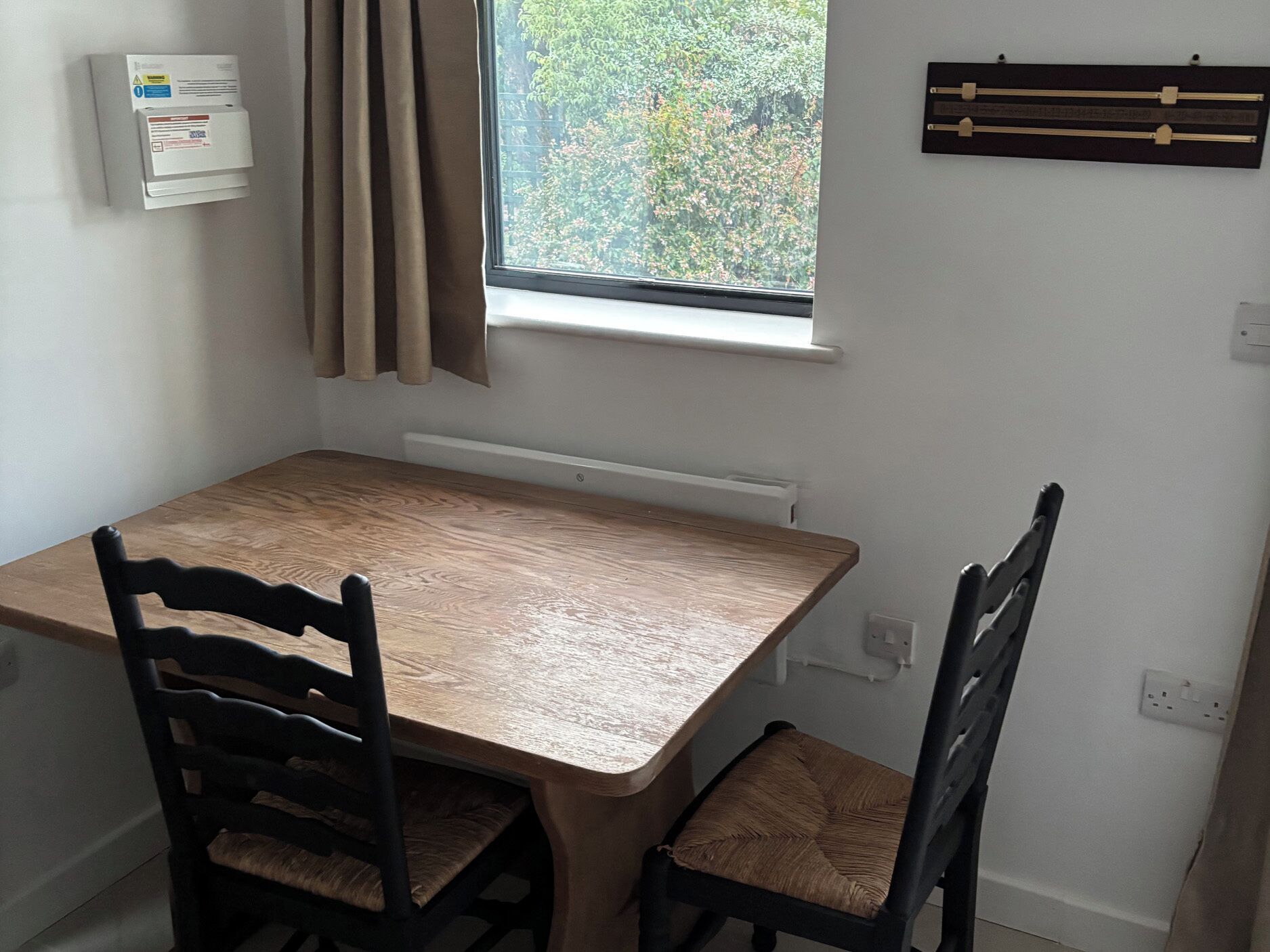 A small dining area with a wooden table and two chairs, a window with curtains, a wall-mounted thermostat, and a power outlet.