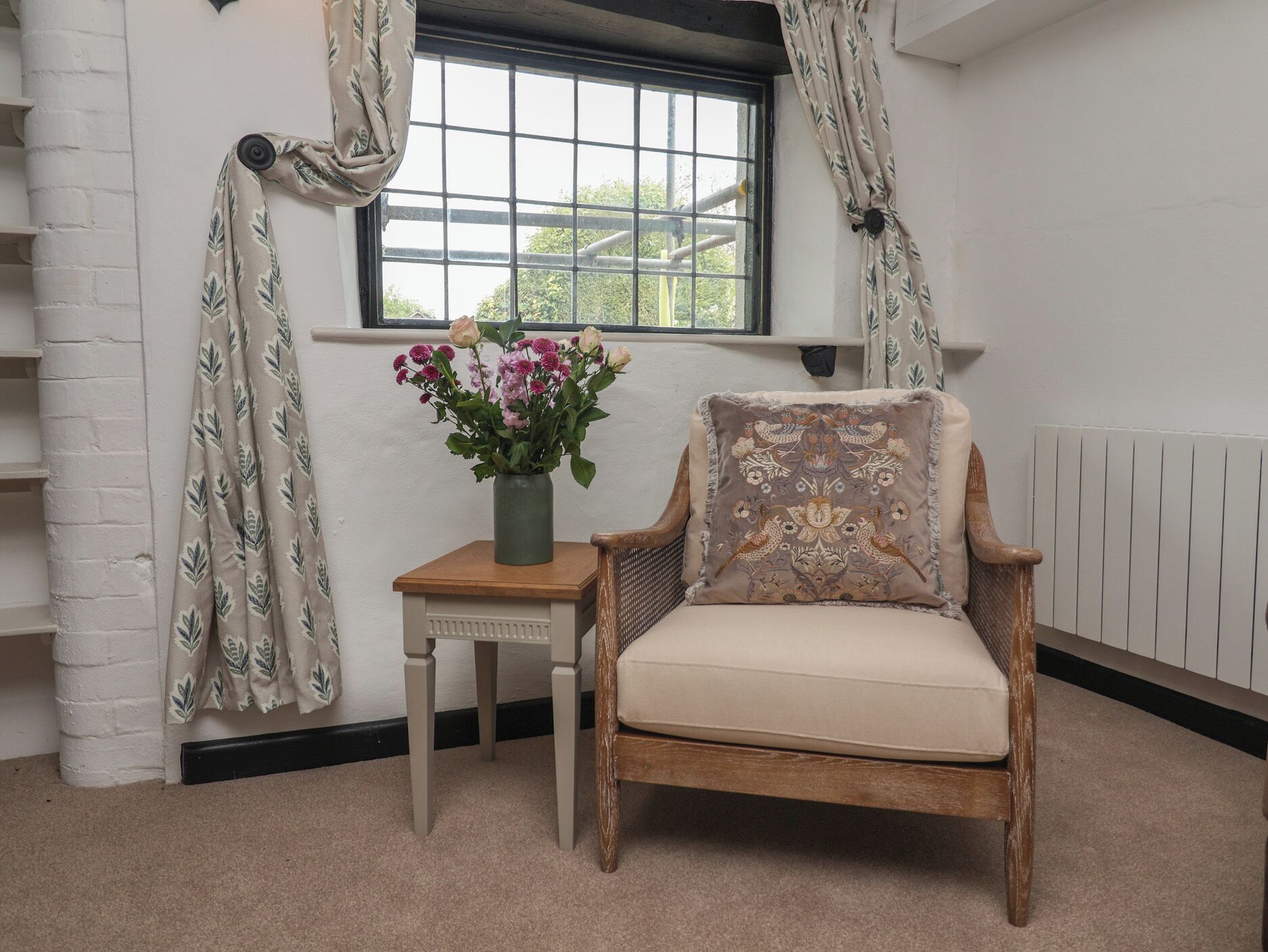 Ein Zimmer mit Fenster, ein Sessel mit Kissen, ein kleiner Tisch mit Blumenvase und eine weiße Backsteinwand.
