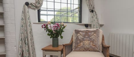 A room with a window, a chair with a cushion, a small table with a vase of flowers, and a white brick wall.