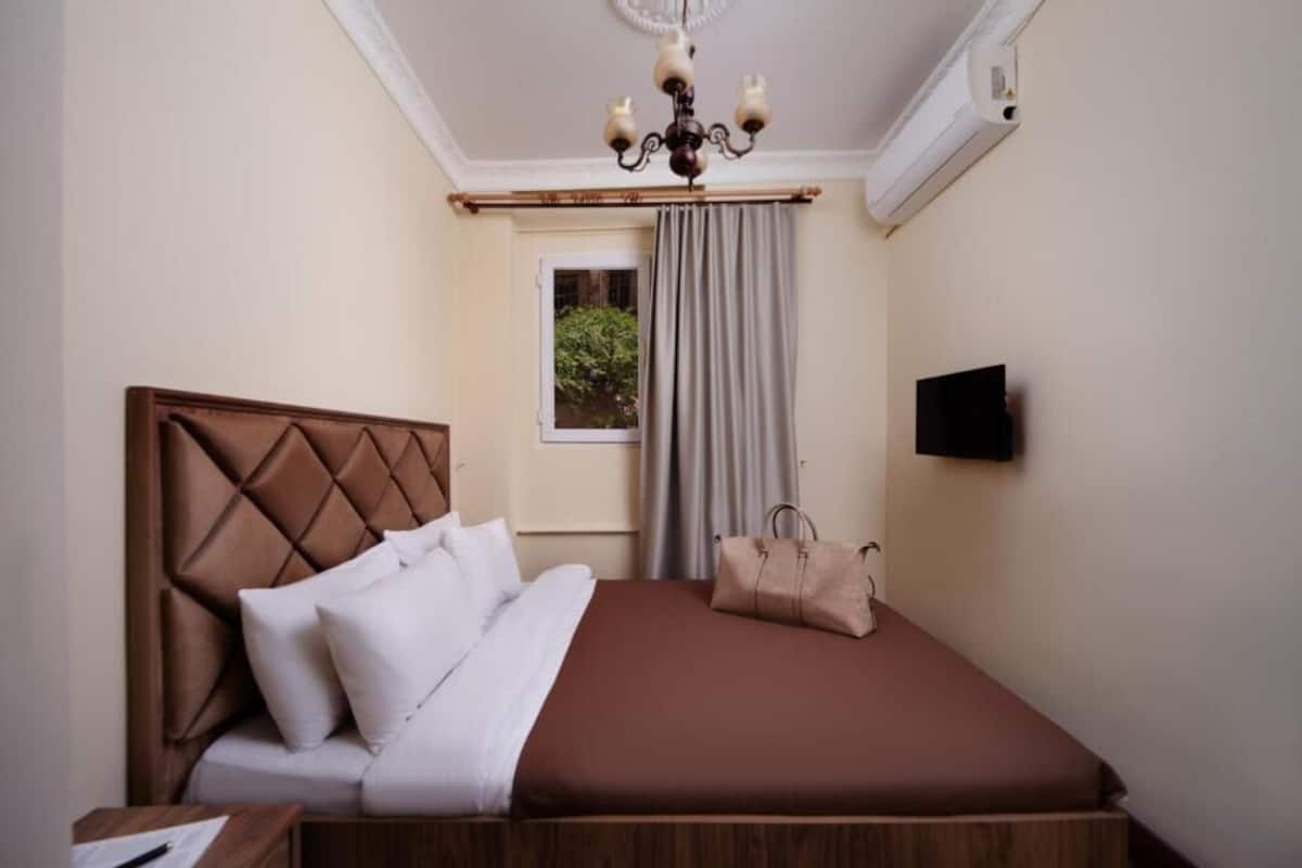 A hotel room with a bed, a nightstand, a TV, a window with curtains, and a chandelier.