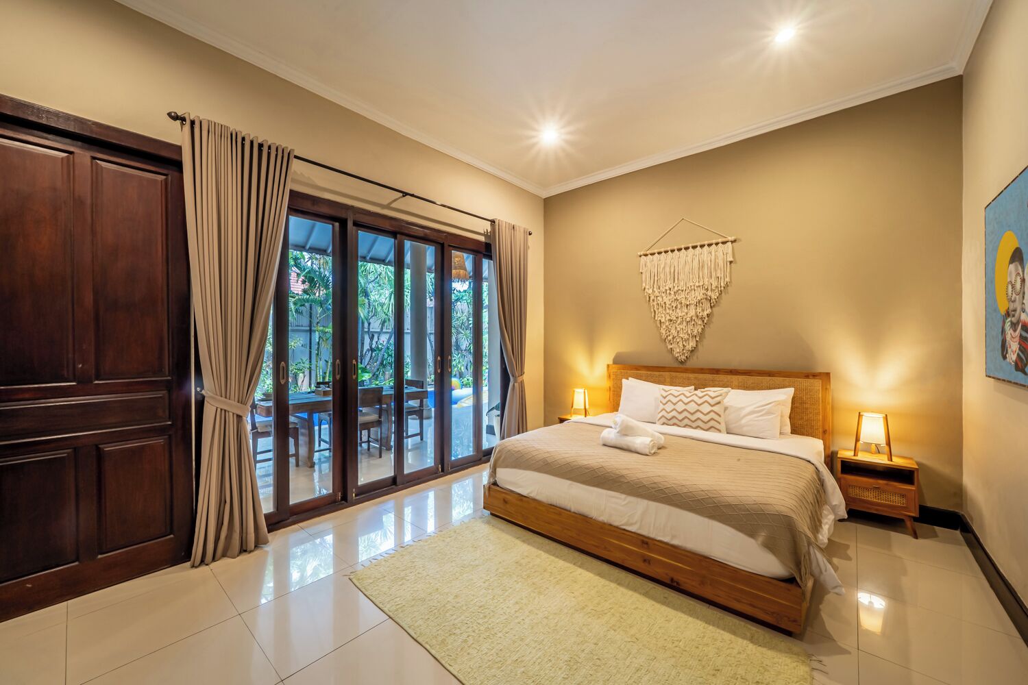A bedroom with a large bed, a wooden headboard, a nightstand, a wall-mounted macrame piece, and a sliding glass door leading to an outdoor area.