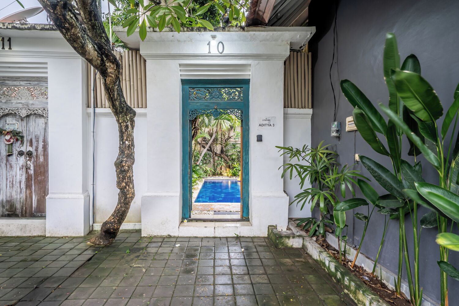 Uma entrada com portão, porta azul, piscina e vegetação exuberante.