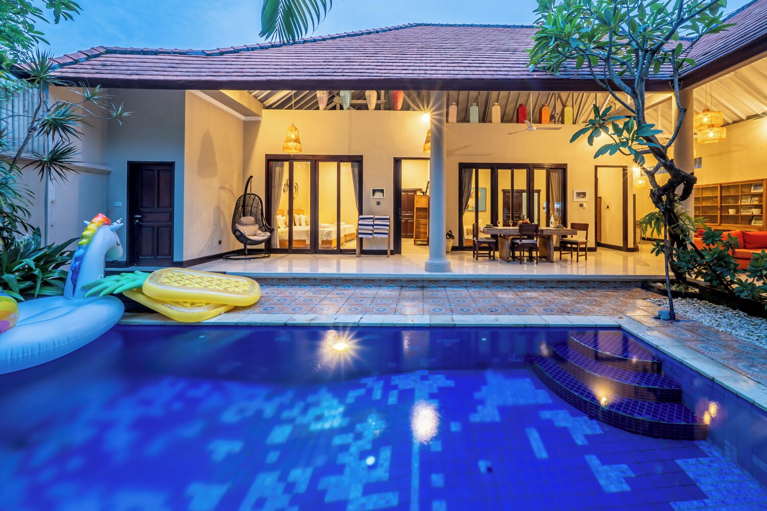 Ein Pool mit bepflasterter Liegefläche, ein Essbereich mit Tisch und Stühlen sowie eine überdachte Außenlounge mit Tisch und Stühlen.
