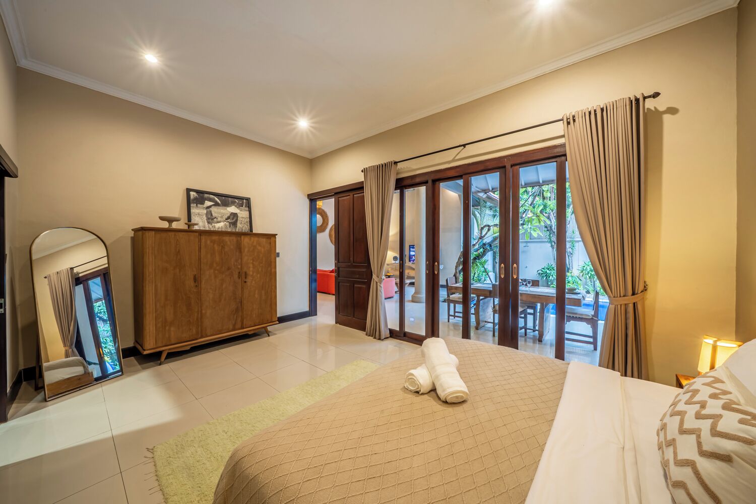 A hotel room with a bed, a wooden wardrobe, a mirror, a window with curtains, and a view of the outside.