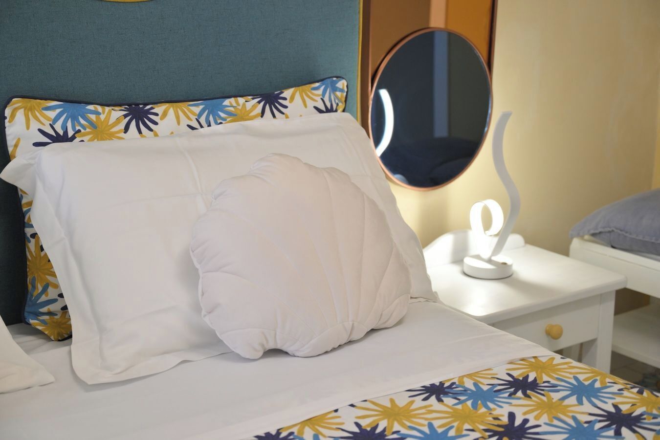 Ein Bett mit einem weißen Kissen und einem Zierkissen, ein Nachttisch mit Spiegel und eine kleine Lampe.