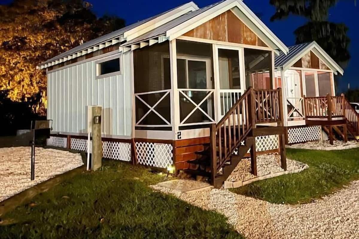 A two-story mobile home with a porch and a small deck, surrounded by gravel and grass.
