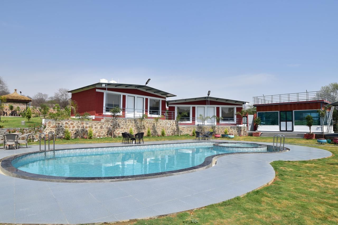 Una piscina con borde de piedra, dos edificios rojos con grandes ventanales y una zona ajardinada con mobiliario de exterior.