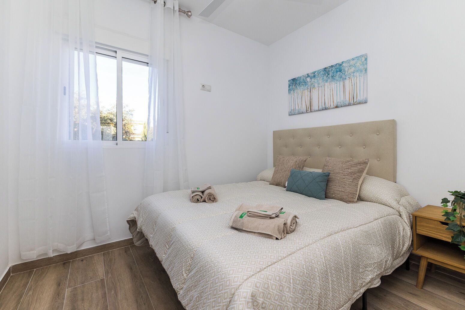 A bedroom with a bed, a nightstand, a painting, and a window with curtains.