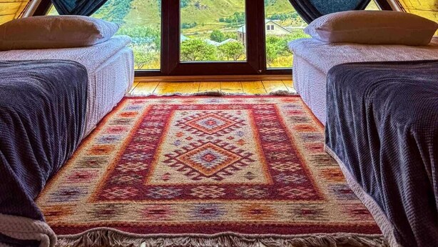 A room with a sloped ceiling, two beds, a large window, and a patterned rug.