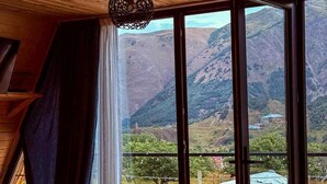 A room with a wooden ceiling, a large window with curtains, a hanging light fixture, and a view of a mountainous landscape.