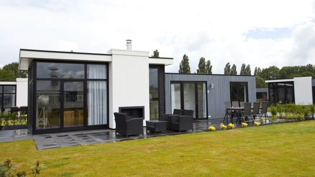 Ett modernt hus med stora glasfönster, en uteplats med utemöbler och en välskött gräsmatta.
