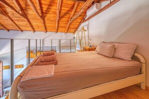 Ein Schlafzimmer mit Holzbalkendecke, einem Bett mit beiger Matratze, Kissen und einem Nachttischchen mit Lampe.