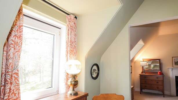 Ein Zimmer mit Fenster, Holzstuhl, Kommode und Lampe.