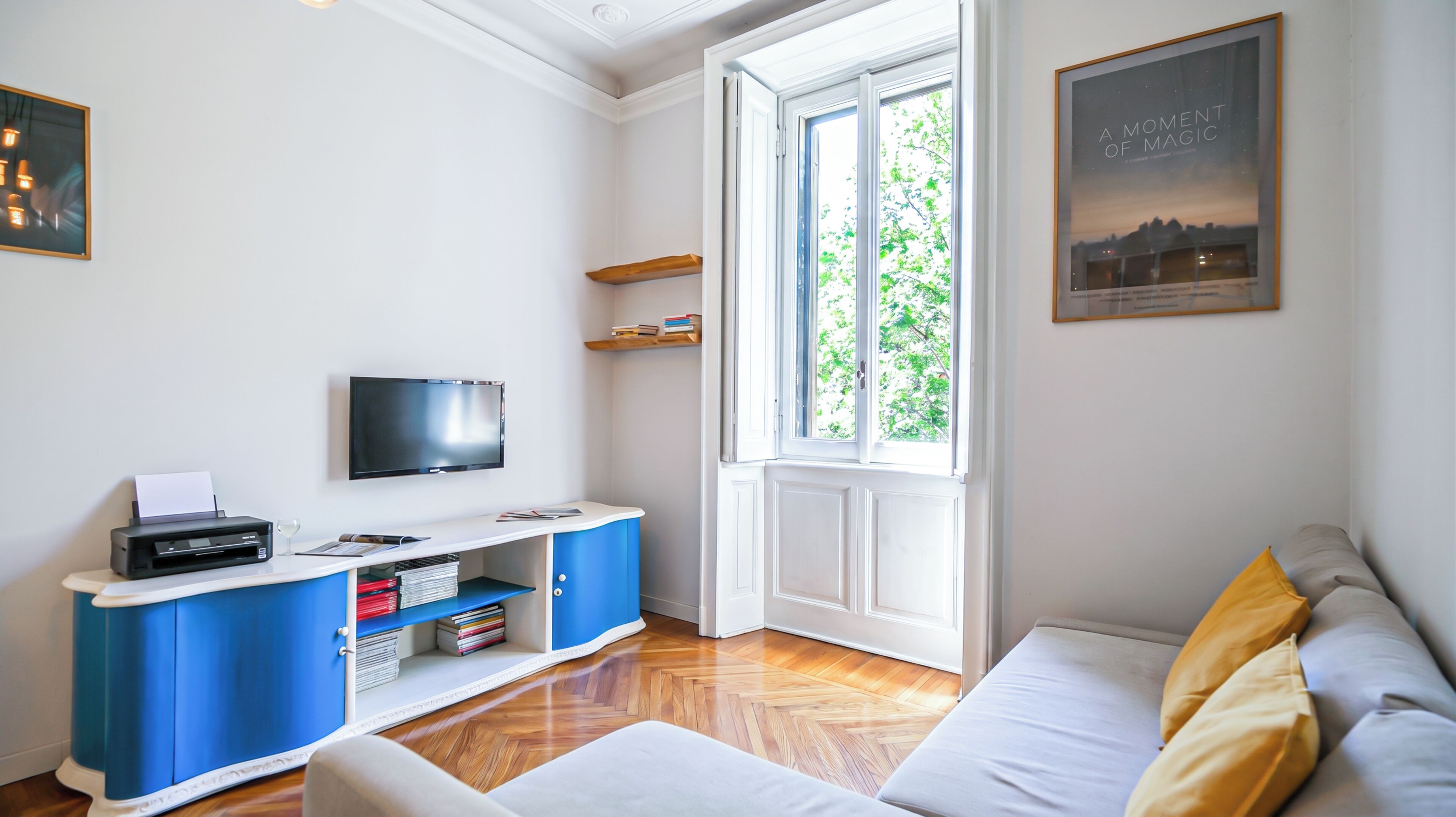 A modern living room with a flat-screen TV, a blue entertainment unit, a sofa, and a window with a view of greenery.