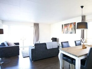 Ein modernes Wohnzimmer mit einer Couch, einem Esstisch und Stühlen. Ein Fenster mit Vorhängen, ein Gemälde an der Wand und eine Lampe sind ebenfalls zu sehen.