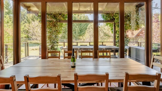 A large wooden dining table with chairs, a bottle of wine, and a view of a garden through large windows.