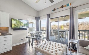 Una sala moderna con televisor de pantalla plana, un comedor con mesa y sillas redondas, un ventanal con cortinas y un balcón con mobiliario exterior.