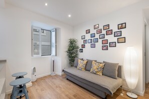 En moderne stue med en grå sofa, et lille bord, en gulvventilator og en væg pyntet med billeder i rammer.