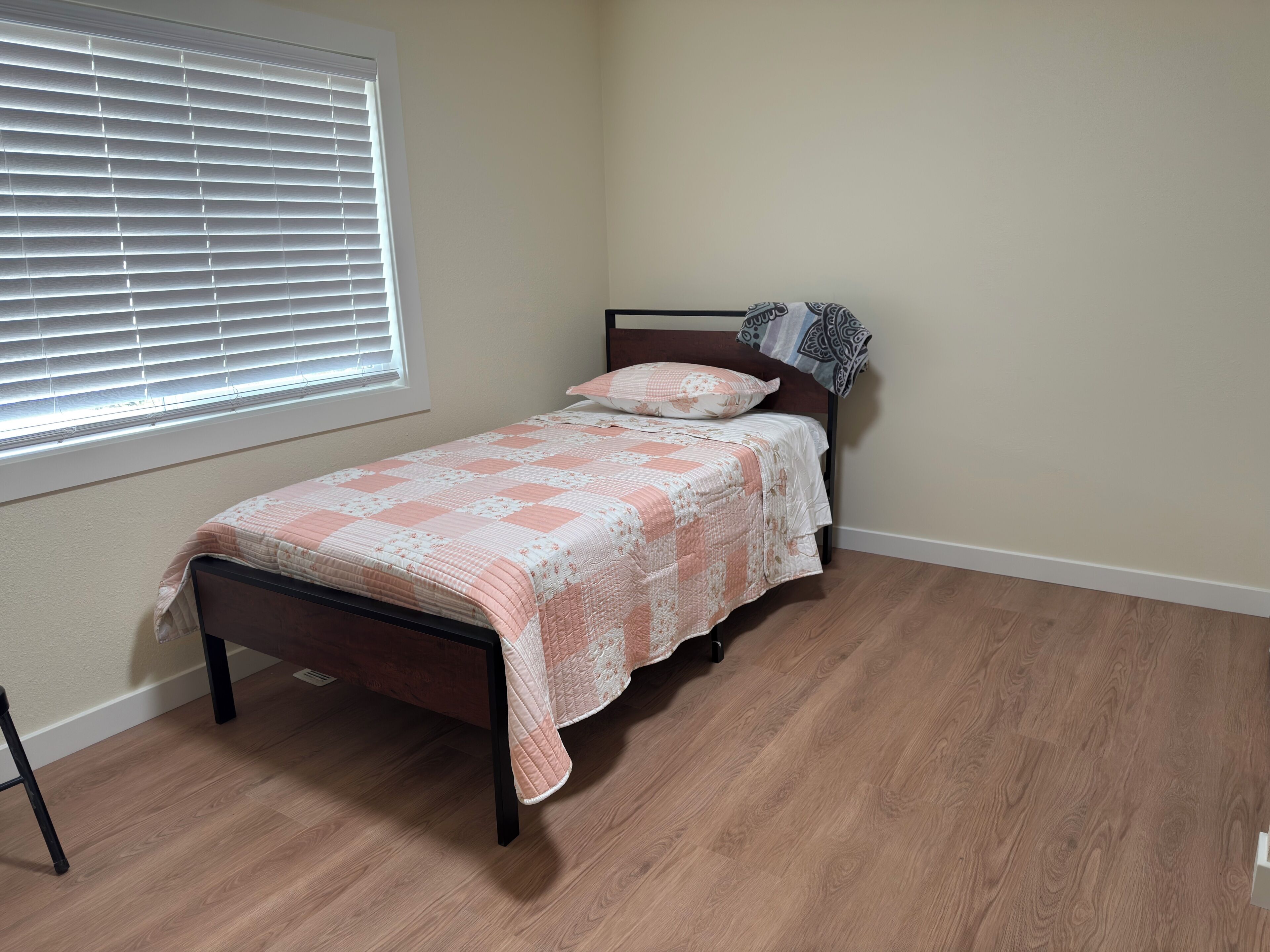 A single bed with a patterned bedspread, a window with blinds, and a wooden floor.